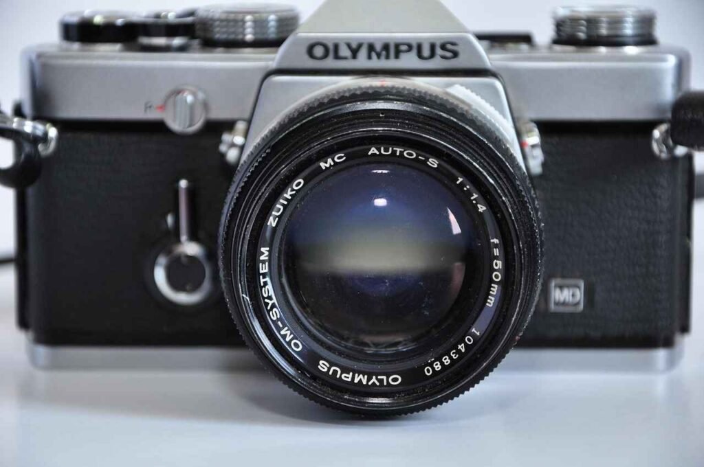 Close-up, frontal view of a vintage Olympus OM-1 film SLR camera with a 50mm f/1.4 lens, sitting on a white surface