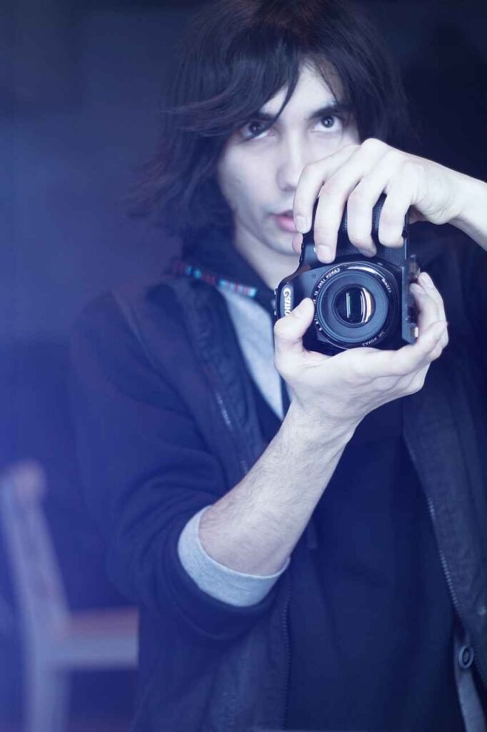 A young person holding an SLR camera up close to their face, looking intently at the viewer through the lens, with dramatic blue-tinted lighting.